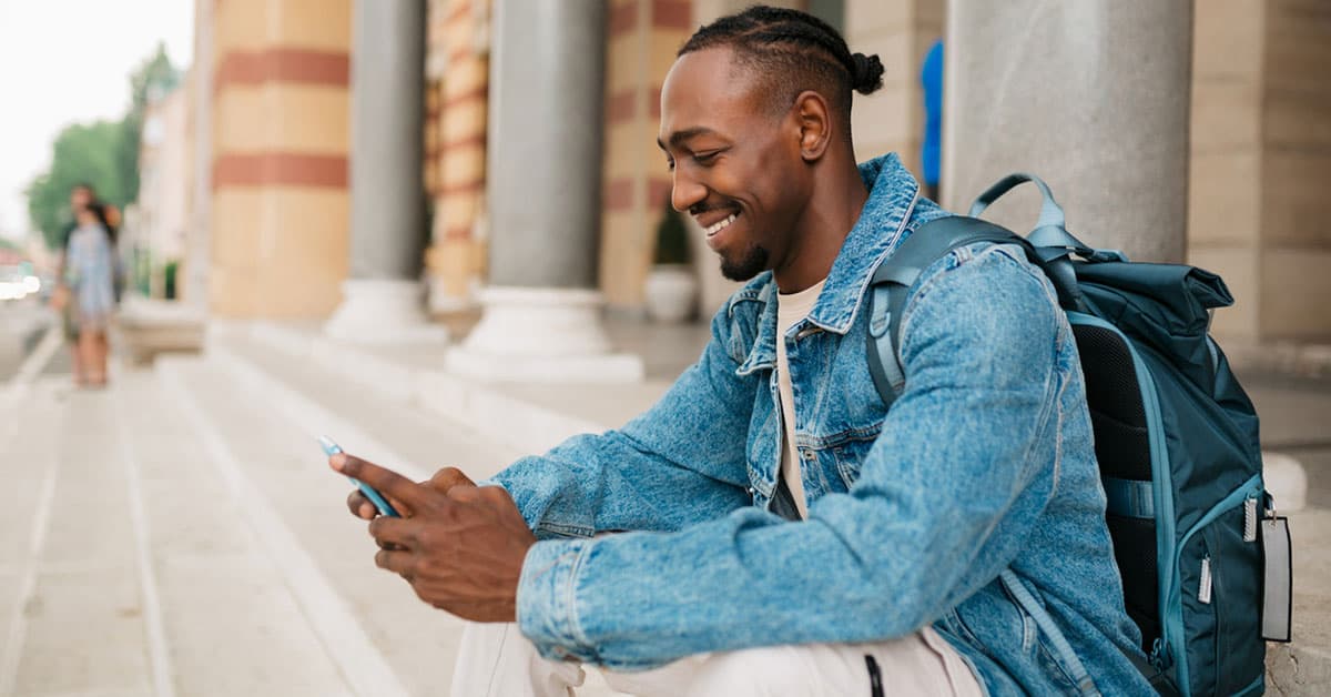 young adult male university student sitting on the steps in front of the university building and using a smart phone